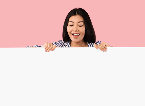 Place For Your Promo. Excited asian woman standing behind white empty board for advertisement over pink background. Lady peeking out big blank advertisement panel, looking down at empty space for text