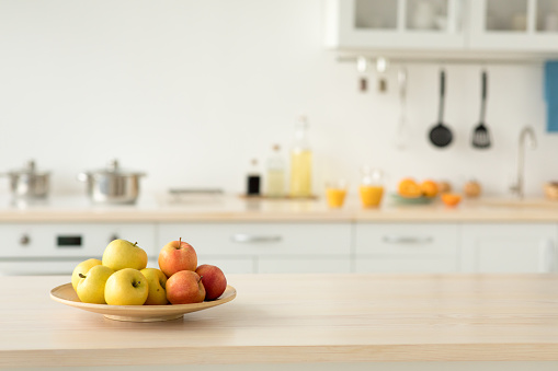 Cozy home kitchen design and comfortable style, ad for rent. Plate with apples on table, bottles of sauces and utensils, orange juice in glasses and fruits, light walls and white furniture, copy space