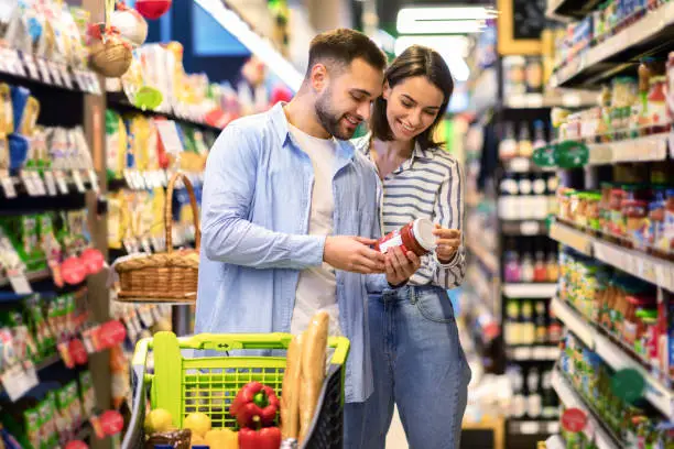 Happy Couple Buying Food In Supermarket, Choosing Products Standing With Trolley Cart Along Aisles And Full Shelves Purchasing Groceries Essentials Together. Smiling Spouses Holding Jar Of Sause