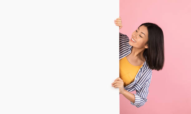 asian lady hiding behind blank white advertising billboard - peeking imagens e fotografias de stock