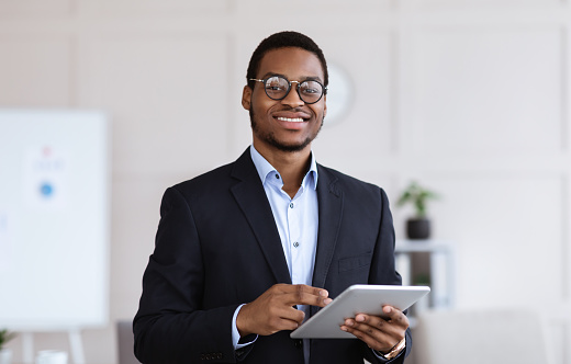 Cheerful young black man manager in glasses holding digital tablet and smiling at camera, office interior, copy space. African american businessman using business application on pad