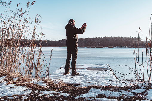 In winter, a man standing on a fishing pier by a frozen lake takes pictures with his phone.