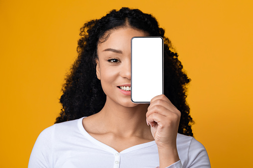 Smiling Black Woman Covering Face With Smartphone With Blank White Screen, Recommending New Mobile App Or Demonstrating Copy Space For Your Design Or Advertisement, Yellow Background, Mockup Image