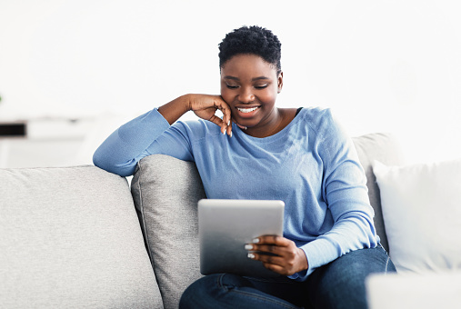 Rest And Relax Concept. Joyful curvy black woman watching movie, using mobile application on pad. Relaxed african american young lady sitting on couch, using digital tablet, home interior