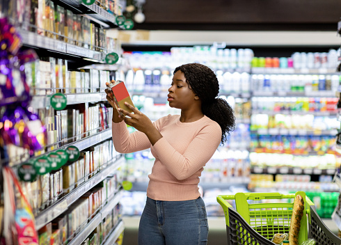 Pretty African American woman choosing groceries at supermarket, shopping for food, buying products for her family. Female consumer purchasing goods at huge mall, walking among shelves