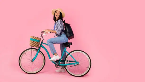 Full length portrait of excited asian woman wearing summer hat and black backpack riding vintage bicycle with wicker basket looking back at free copy space isolated on pink studio background
