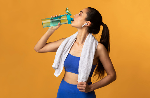 Fitness And Healthy Lifestyle Concept. Portrait of sporty young woman drinking mineral water from bottle at studio after workout at gym, wearing earbuds, banner. Wellness And Training