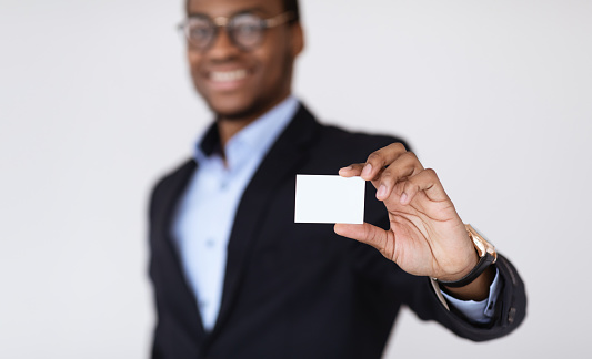 Business Card Mock-Up. Smiling black businessman holding blank card for clients or business partners, selective focus on empty white card, grey studio background, closeup, copy space