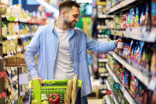 Everyday Shopping Concept. Smiling Young Man Pushing Trolley Cart Between Aisles In Grocery Store. Positive Guy Buying Food, Taking Products From Shelf. Consumerism And Lifestyle