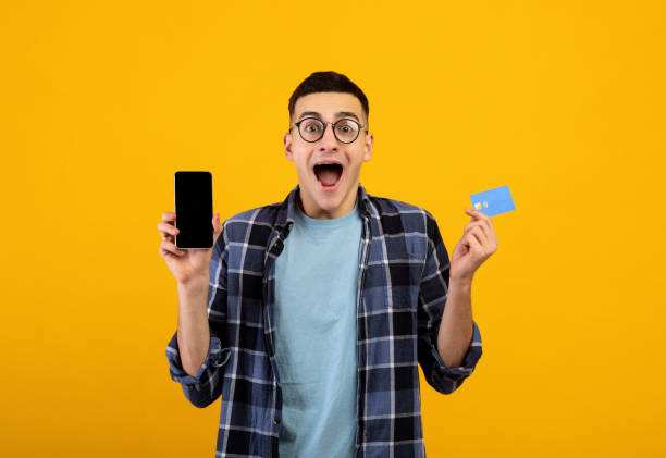 istock Excited millennial guy holding credit card and smartphone with empty screen on orange background, space for design 1305348199