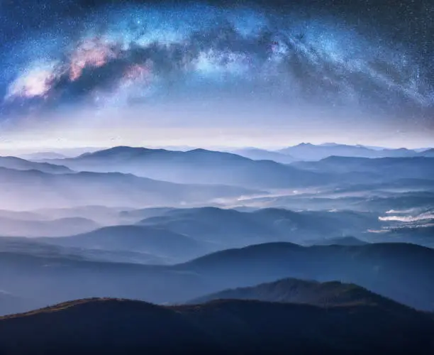 Photo of Milky Way arch over the mountains in fog at starry night in summer. Landscape with blue sky with stars, arched Milky Way, trees on the foggy hills, mountain peaks. Space and galaxy. Aerial view