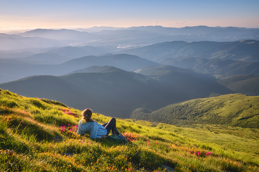 Young woman on the mountain peak with green grass and pink flowers looking at beautiful mountains in fog at sunset in summer. Colorful landscape with lying girl, forest, hills, sky. Travel and tourism