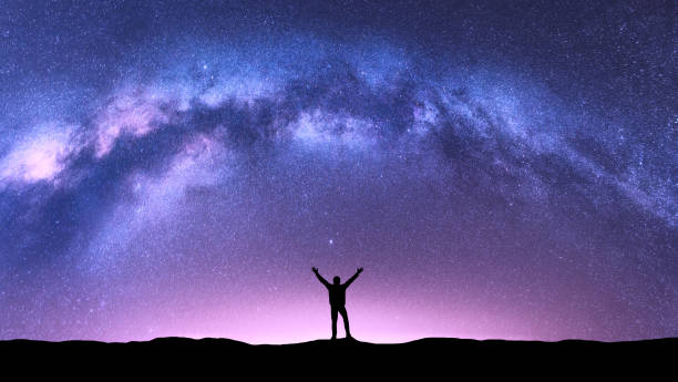 Photo of Arched Milky Way and happy man at night. Silhouette of guy with raised up arm on the hill, purple sky with stars, pink light in summer. Galaxy. Space background. Landscape with milky way arch. Travel