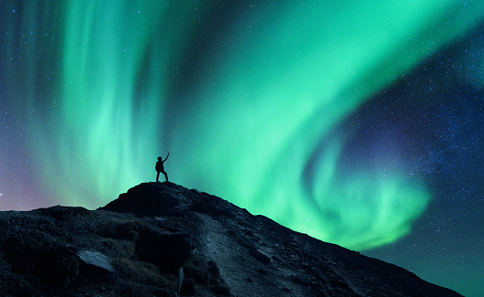 Northern lights and silhouette of standing man with raised up arms on the mountain in Norway. Aurora borealis and happy man. Sky with stars and green polar lights. Night landscape with aurora. Concept