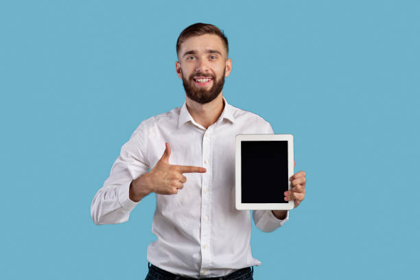 istock Young businessman pointing at tablet with empty screen on blue background, mockup for application or website design 1305348726