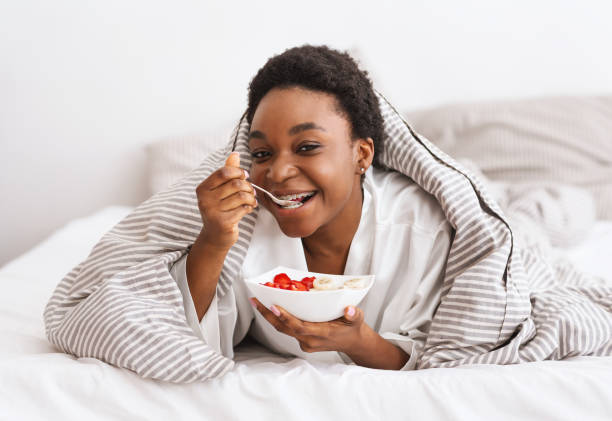 Healthy breakfast at home and body care during covid-19 lockdown Healthy breakfast at home and body care during covid-19 lockdown. Smiling young african american lady lies on bed under blanket with plate, eats porridge with fruits, in bedroom interior, copy space bed breakfast pictures stock pictures, royalty-free photos & images