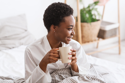 Coffee for breakfast, favorite latte, vitality and energy in morning. Cheerful young cute african american female sitting on bed with cup of hot drink, in bedroom interior before work, copy space