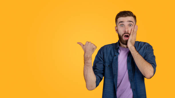 istock Promotion concept. Shocked hipster guy pointing aside at free space, recommending something on orange studio background 1305350051
