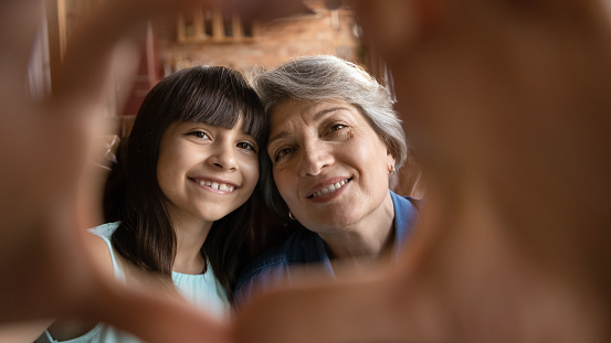 Close up portrait of smiling mature Hispanic grandmother and little 7s granddaughter have fun make selfie together. Happy senior Latino grandparent and small girl child make heart love hand gesture.