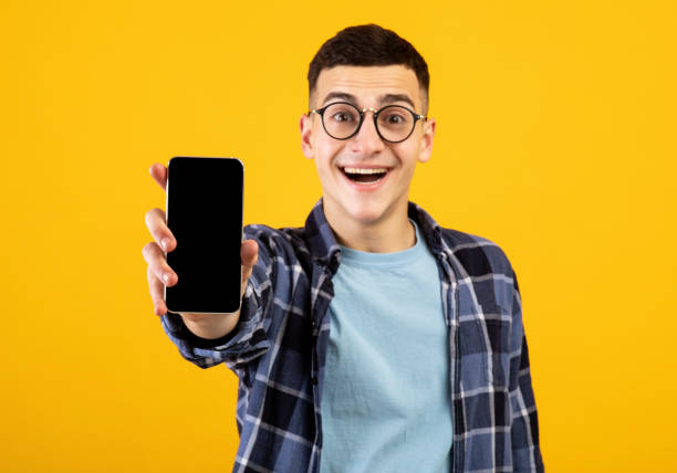 istock Happy young man showing mobile phone with empty screen on orange background, mockup for mobile app or website design 1305351196