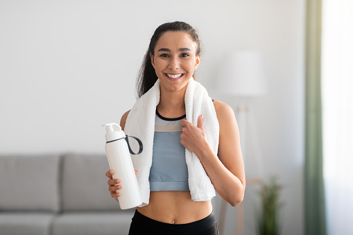Cheerful brunette woman in sportswear having break after training at home, holding bottle with water or protein drink and carrying towel on her shoulders, smiling at camera, copy space