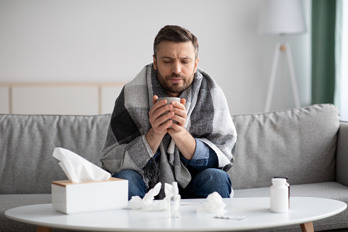 Middle-aged bearded man covered in blanket feeling sick, holding mug with hot drink, having napkins on coffee table, home interior, copy space. Cold, flu or coronavirus infection concept