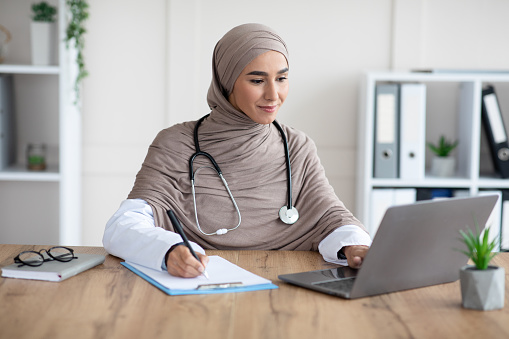 Young woman doctor having online appointment, sitting in office, using laptop and taking notes, copy space. Attractive muslim lady physician working with computer, clinic interior, remote healthcare