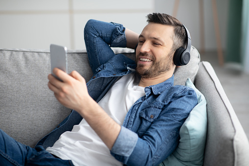 Time to relax. Handsome middle-aged bearded man in wireless headphones listening to music online on smartphone, copy space, closeup. Happy man in headset watching videos on mobile phone