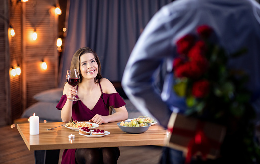 Affectionate Man Prepared Romantic Surprise For His Girlfriend, Holding Roses And Gift Behind Back, Greeting With Valentine's Or International Women's Day, Loving Couple Having Date In Restaurant