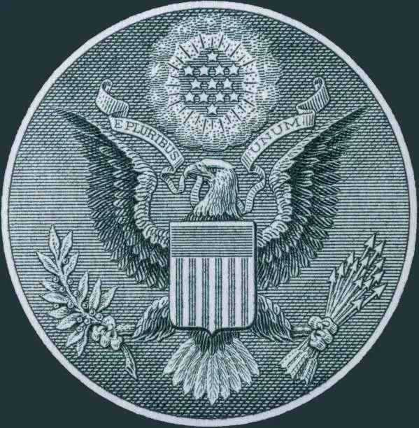 Photo of Great Seal of the United States obverse from the back of a one dollar bill. The Bald Eagle carries a ribbon with E pluribus unum, thirteen arrows and an olive branch, with a glory Or with 13 stars.