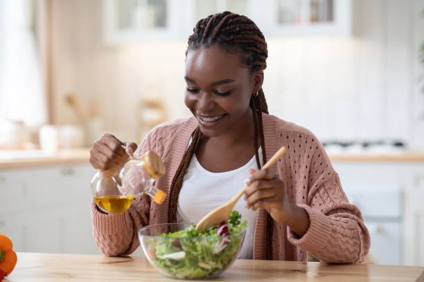 femme afro-américain de sourire faisant cuire la salade végétale dans la cuisine, ajoutant l’huile d’olive - black olive photos et images de collection