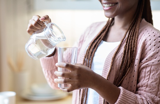 Smiling African American Woman Pouring Water From Jug To Glass. Unrecognizable Black Female Drinking Healthy Liquid In Kitchen, Enjoying Refreshing Drink, Cropped Image, Closeup Shot