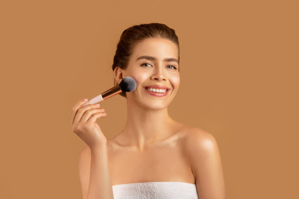 istock Pretty young lady using cosmetic brush to apply decorative makeup on her face, brown studio background 1305354308