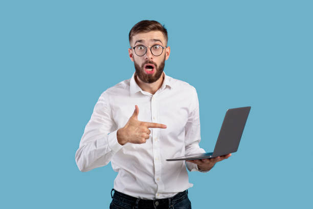 istock Shocked young man pointing at laptop computer with open mouth, surprised over web content on blue studio background 1305354400