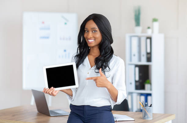 istock Smiling black business lady pointing at tablet computer with blank screen at office, mockup for app or website design 1305354449