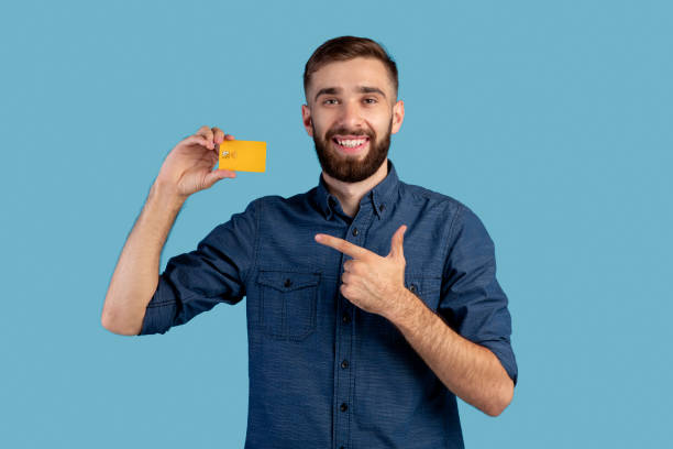Joyful millennial man pointing at credit card on blue studio background. Electronic money concept