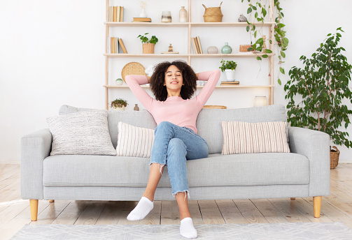 Facial emotions, rest at home, design blog and relaxation after work. Happy calm millennial african american woman with closed eyes sits on sofa with hands behind head in cozy living room interior