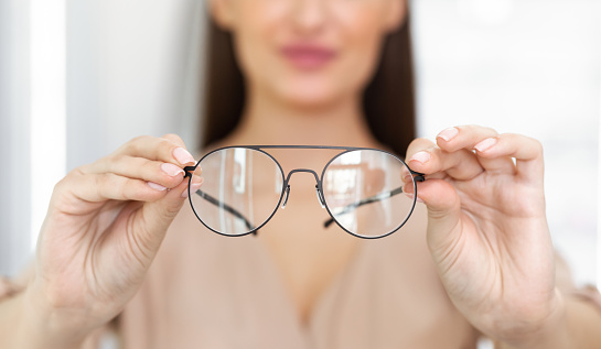 Eyesight And Vision Concept. Closeup of unrecognizable woman showing new eyeglasses to camera, standing at optics store, blurred background, selective focus on eyewear. Lady offering specs