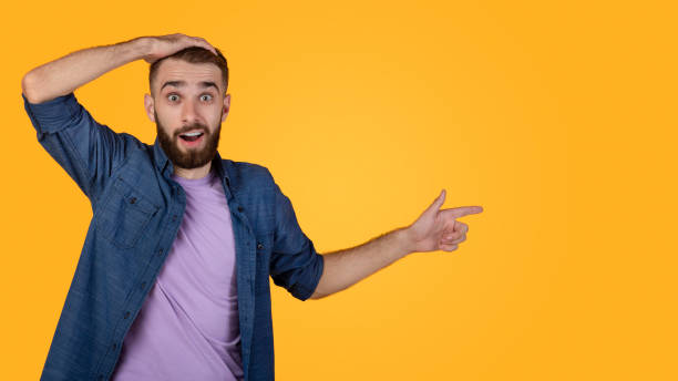 istock Shocked young man pointing aside at blank space on orange studio background, banner design 1305356515