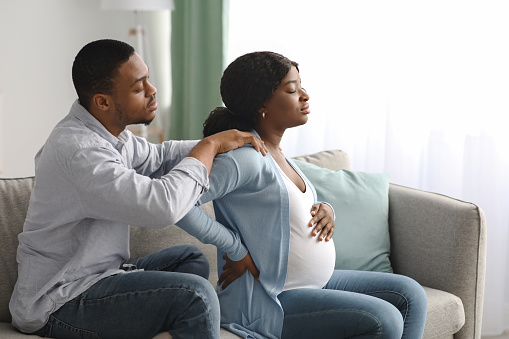 Black pregnant woman suffering from back pain at home, attentive spouse comforting her. African expecting lady having childbirth labor while sitting with husband on sofa at home, copy space