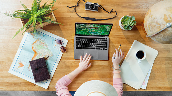 Top view of unrecognizable young woman with laptop and maps planning vacation trip holiday, desktop travel concept.
