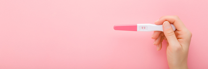 Mujer adulta joven sosteniendo a mano la prueba de embarazo con dos rayas sobre fondo de mesa rosa claro. Color pastel. Resultado positivo. Closeup. Disparo de punto de vista. Amplia pancarta. Lugar vacío para el texto. Vista superior. photo