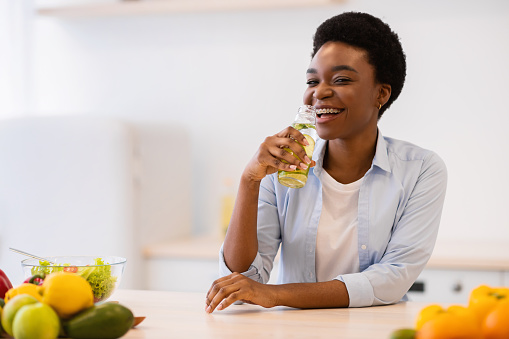 Healthy Hydration. Cheerful Black Lady Drinking Infused Water For Refreshment Posing With Bottle Sitting In Modern Kitchen At Home. Stay Hydrated. Nutrition For Slimming And Weight Loss Concept