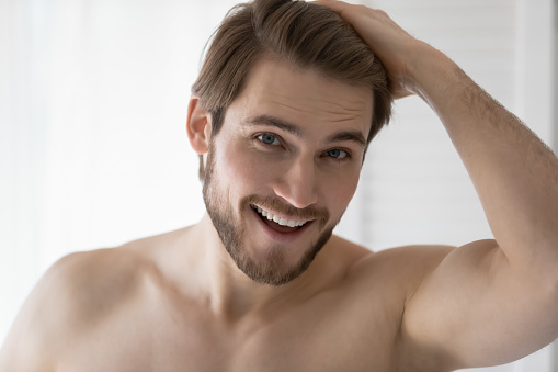 Head shot close up smiling handsome man touching hair, feeling confidence after morning hygienic procedures. Happy 30s caucasian guy looking at camera, showing good mood after refreshment routine.
