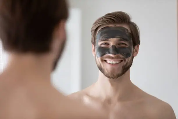 Head shot close up mirror reflection smiling handsome young man grooming himself, applying black charcoal mask on face, feeling satisfied with domestic cleansing skincare procedures in bathroom.