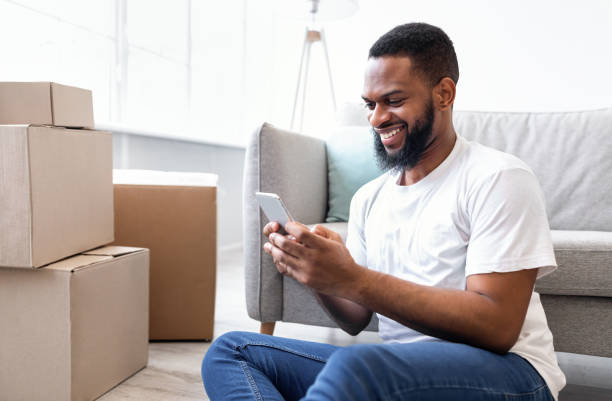 istock Happy Black Man Using Phone Sitting Among Moving Boxes Indoor 1305364254