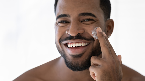 Close up head shot happy laughing young 30s african american handsome man applying moisturizing cream on cheeks, feeling excited of using professional cosmetics in domestic skincare procedure.