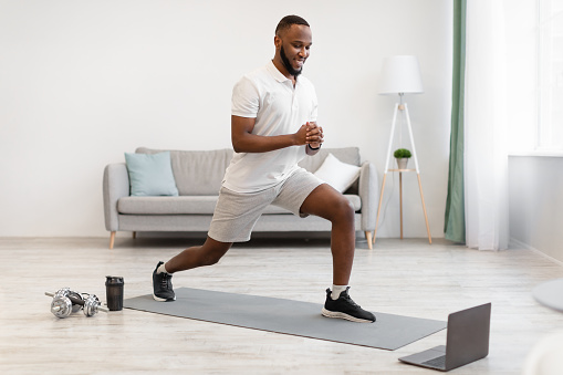 Online Workout. African American Guy Doing Forward Lunge Exercise Watching Fitness Video At Laptop Training At Home. Distant Digital Gym Website, Modern Sporty Lifestyle Concept