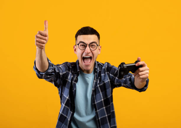 istock Millennial guy with joystick showing thumb up and shouting in excitement, recommending video game on orange background 1305364767