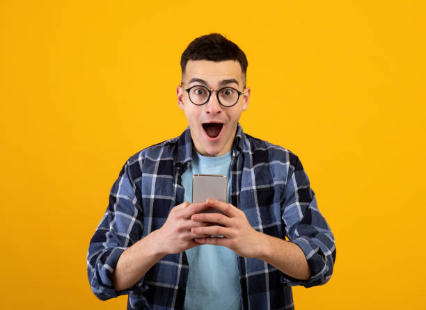 istock Handsome young guy in glasses looking at his smartphone in shock on orange studio background 1305364827